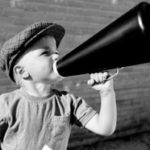 Black and white image of little boy wearing a hat shouting in a megaphone