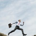 Man holding a briefcase leaping across two cliffs