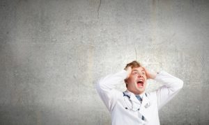 Male doctor holding head while screaming in frustration against gray background