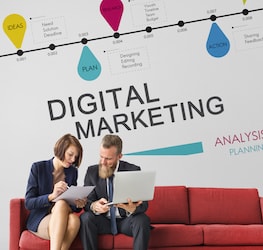 Two professionals sitting on a couch discussing paperwork in front of "Digital Marketing" wall