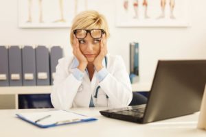 Female doctor wearing glasses, sitting in front of laptop with an expression of frustration