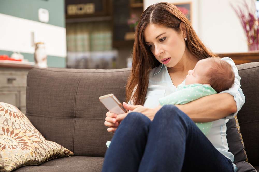 Woman sitting on a couch, holding a baby, looking intently at cellphone