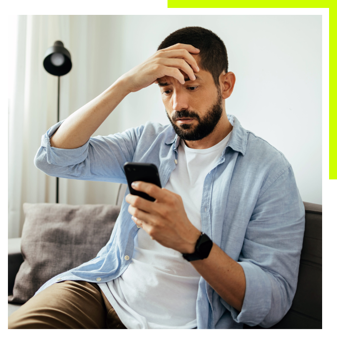 A man sitting on a couch, looking at his smartphone with a stressed and worried expression, his hand resting on his forehead.