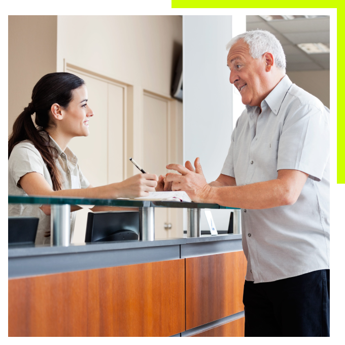 A smiling female receptionist assisting a senior man at a front desk check-in counter.