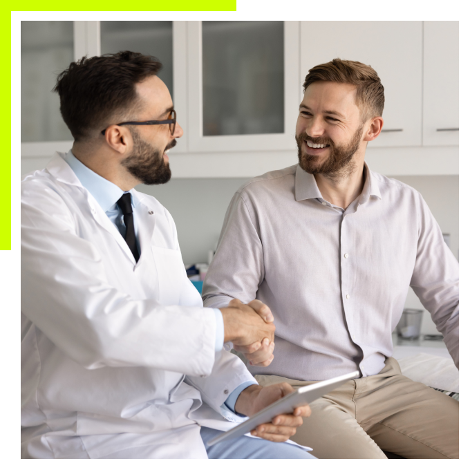 A smiling doctor in a white lab coat shaking hands with a male patient during a consultation in a bright medical office.