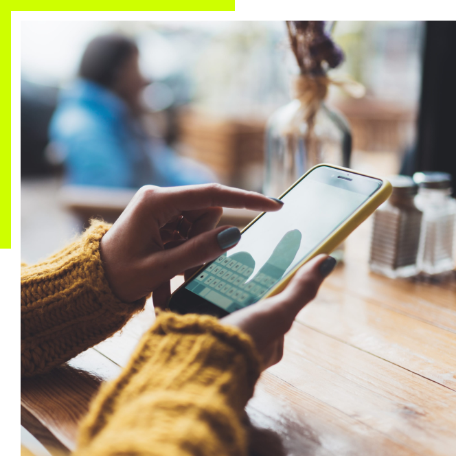 A close-up of a person in a yellow sweater using a smartphone at a wooden cafe table.